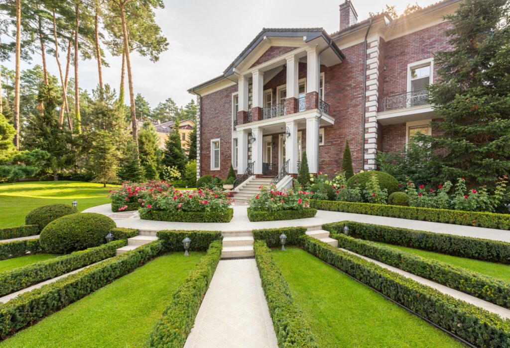 Luxurious brick mansion surrounded by lush gardens and trimmed hedges on a sunny day.