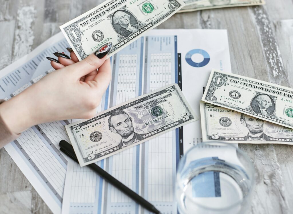 Close-up of a hand holding a dollar bill over financial documents and charts on a wooden table.