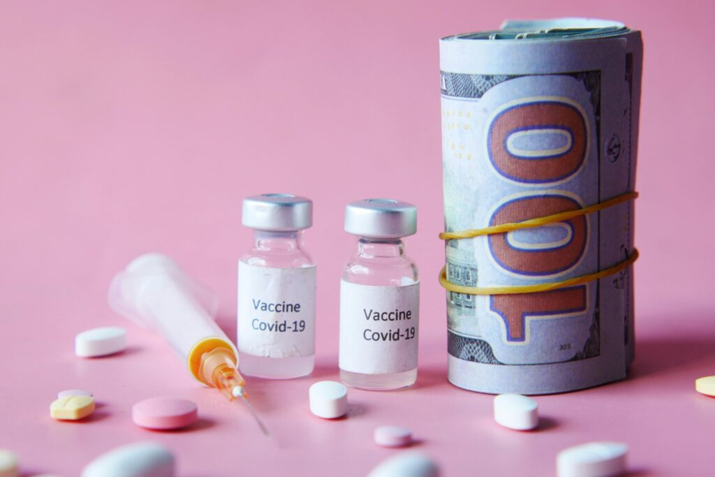 Covid-19 vaccine vials, US currency, and pills on a pink background, representing healthcare costs.