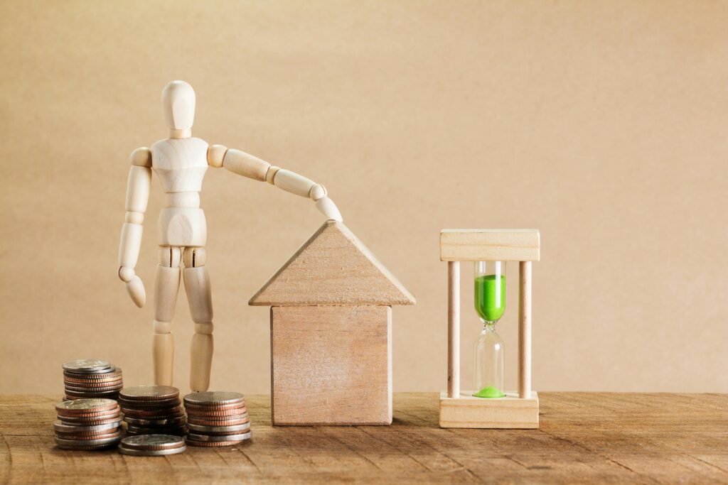 Wooden mannequin with house model, coins, and hourglass symbolizing financial planning.