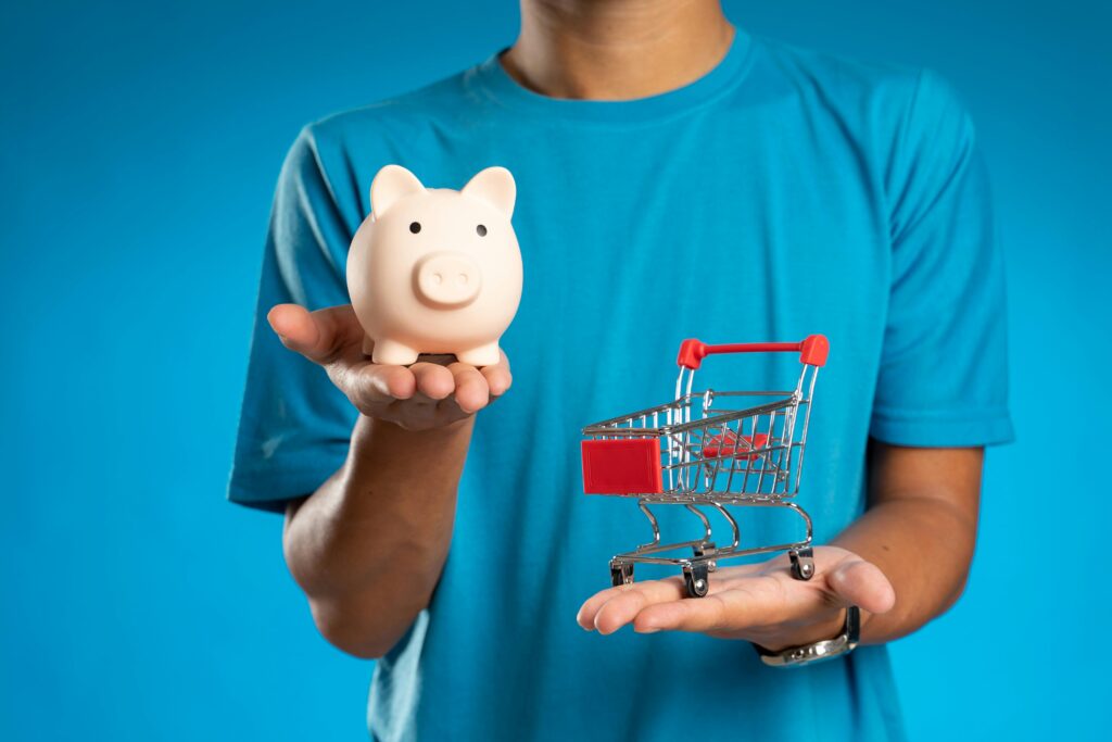 Man in blue shirt holding piggy bank and mini shopping cart, symbolizing savings versus spending.