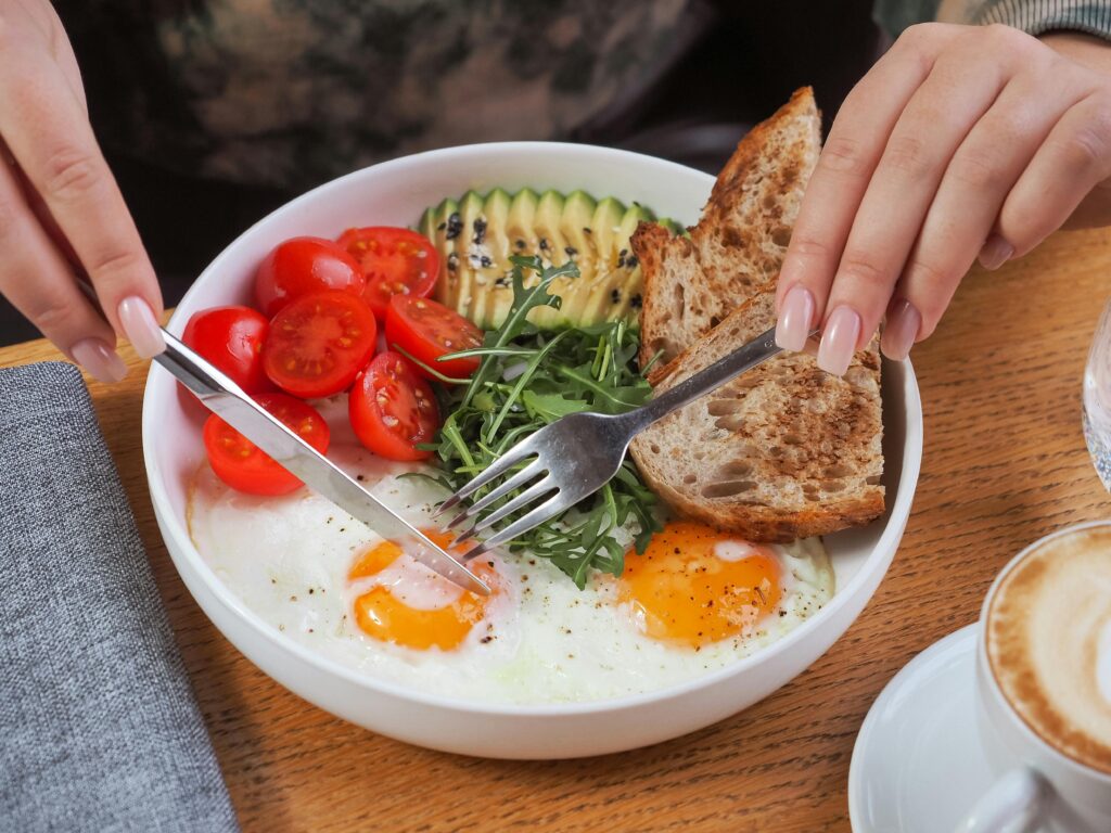 Close-up of a breakfast bowl with fried eggs, avocado, tomatoes, and bread, served with cappuccino.