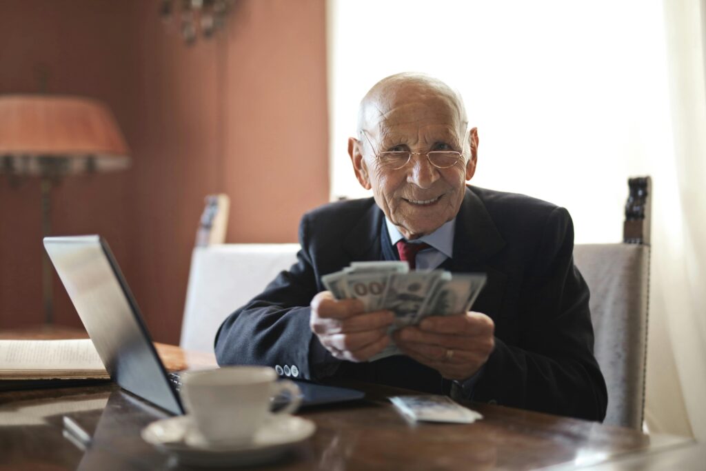 Positive senior businessman in formal suit and eyeglasses holding money in hands while sitting at table near laptop and looking at camera