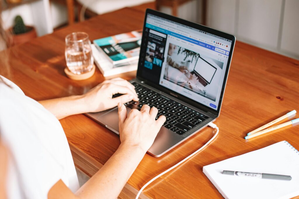 A person working remotely on a laptop at a wooden desk with stationery and a notepad.